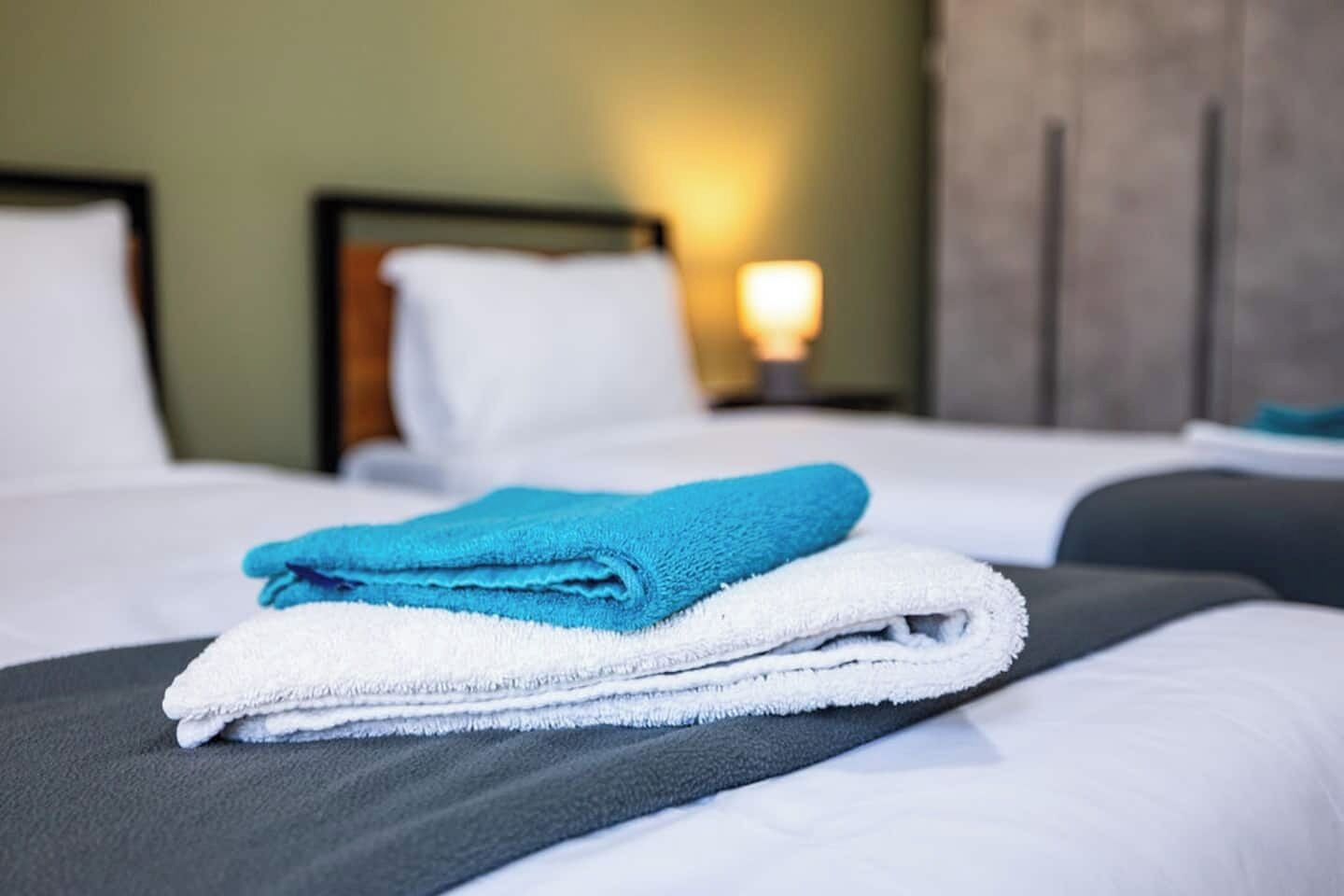 Una cama bien hecha con una toalla blanca y azul doblada, un cabecero de madera y una lámpara fijada a la pared.