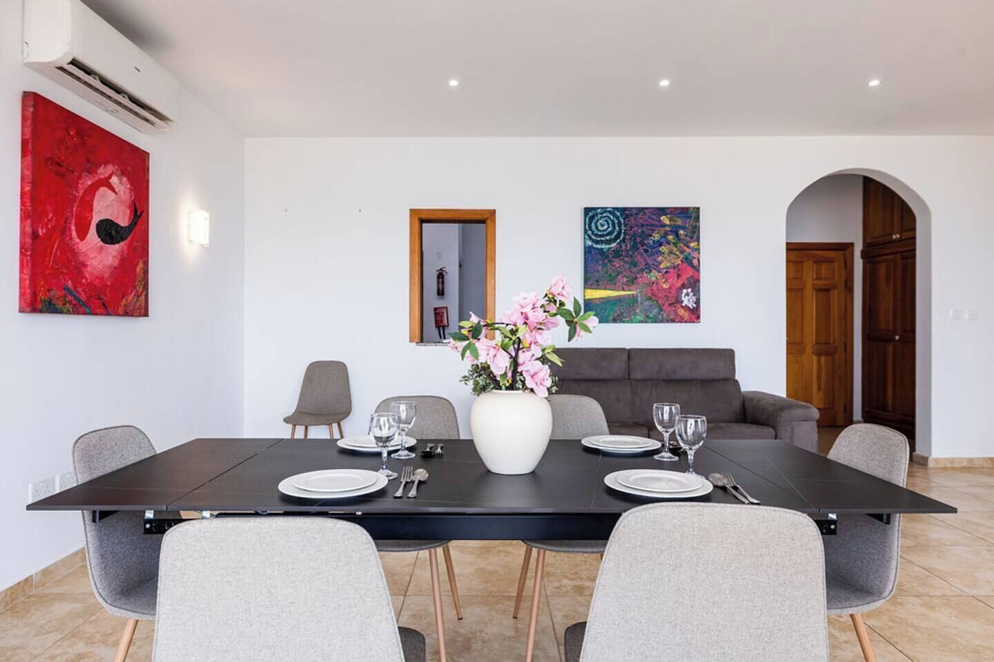 Ein Essbereich mit einem rechteckigen Tisch, gedeckt für sechs Personen, umgeben von grauen Stühlen. Auf dem Tisch steht eine große, weiße Vase mit rosa Blumen.