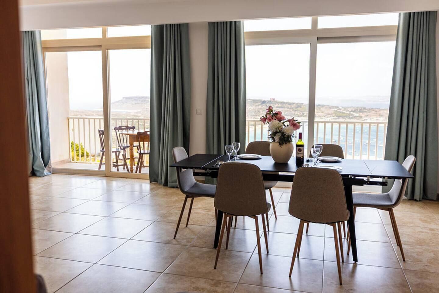 A dining area with a table set for four, a vase with flowers, and a view of the sea through large windows.