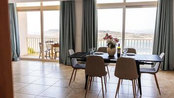 A dining area with a table set for four, a vase with flowers, and a view of the sea through large windows.