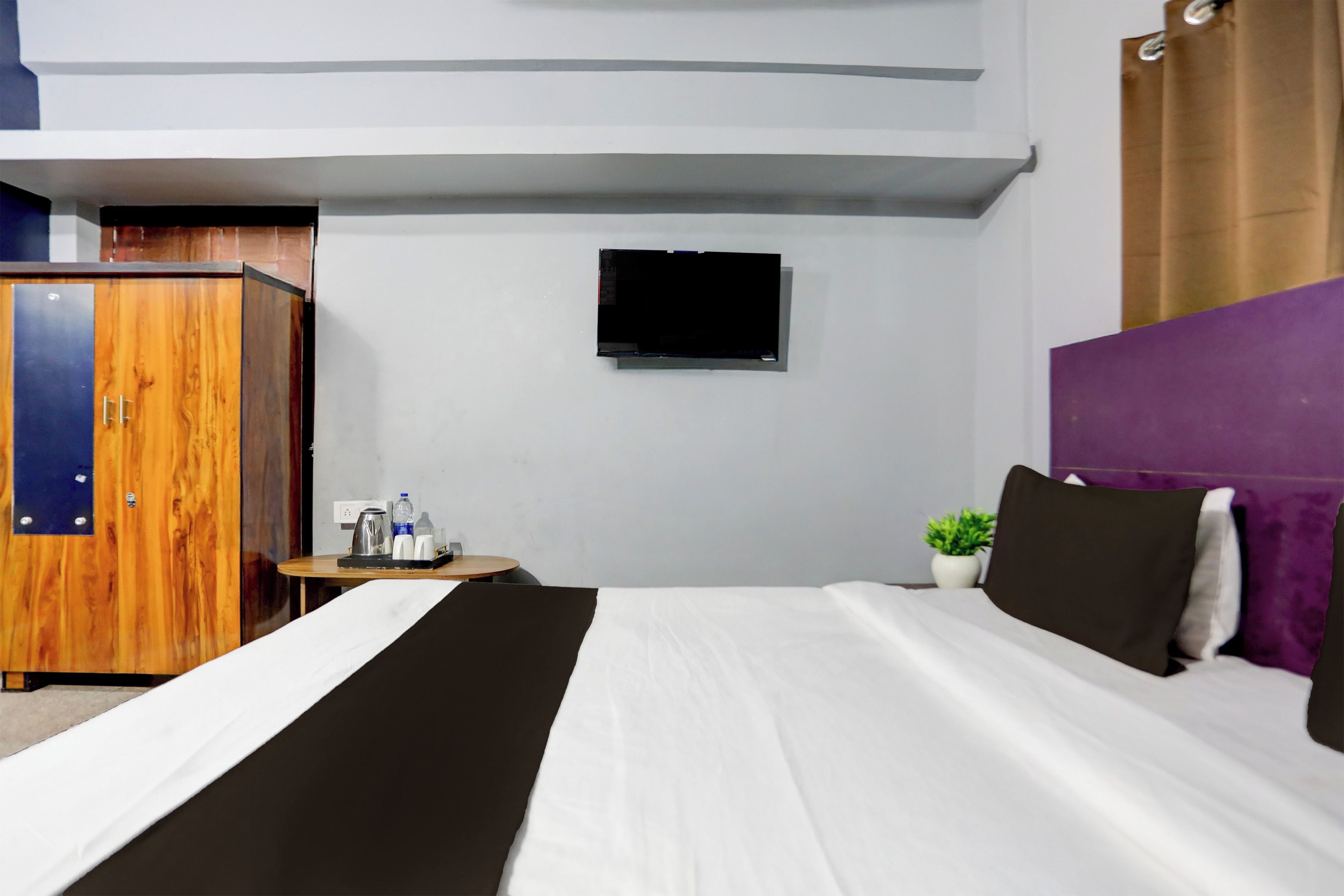 Ein Hotelzimmer mit einem Bett, einem an der Wand befestigten Fernseher, einem Holzschrank, einem kleinen Tisch mit Wasserkocher und Tassen sowie einer Zimmerpflanze.
