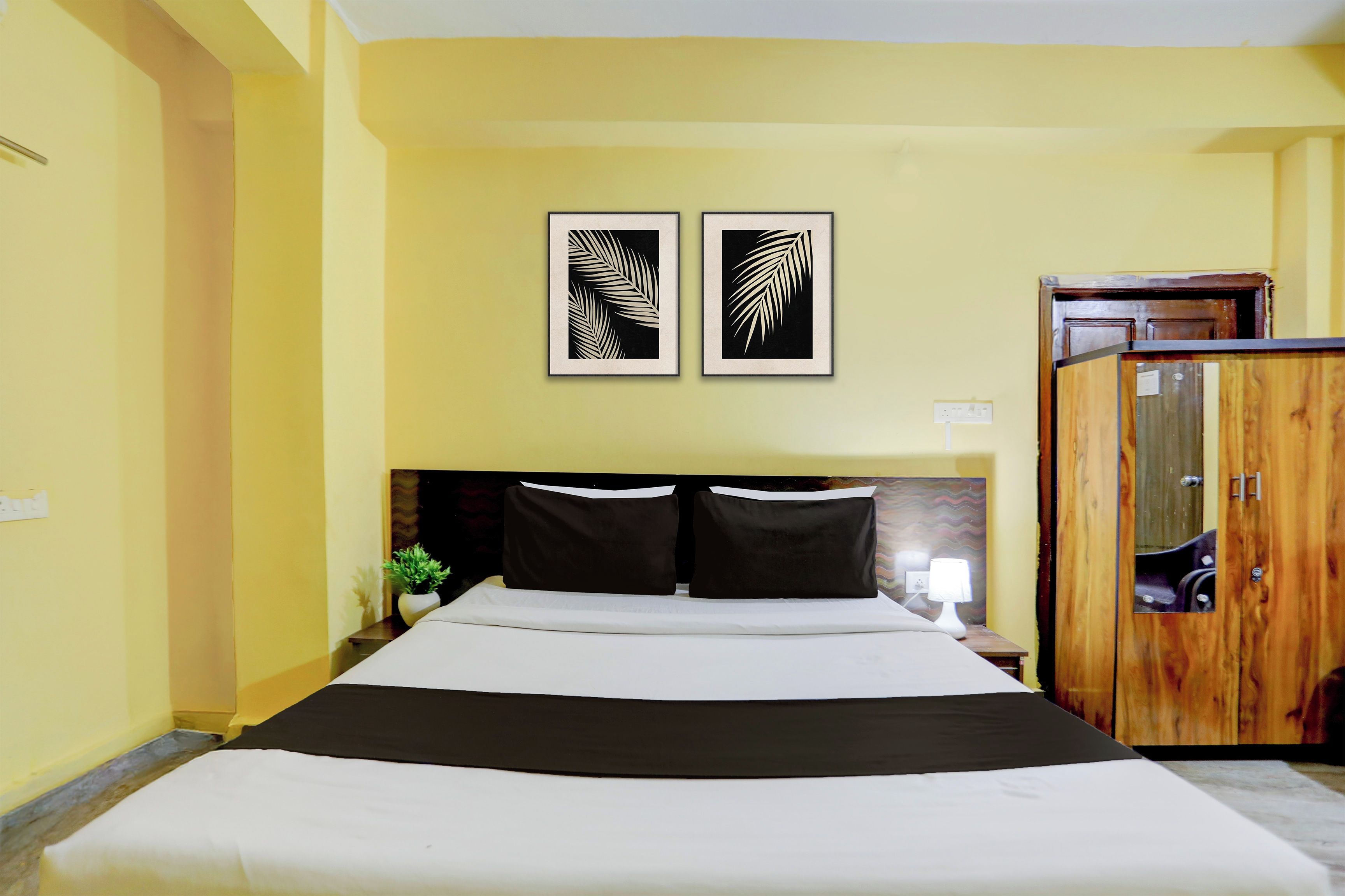 A hotel room with a bed, two framed leaf wall art, a wooden wardrobe, and a nightstand with a lamp.
