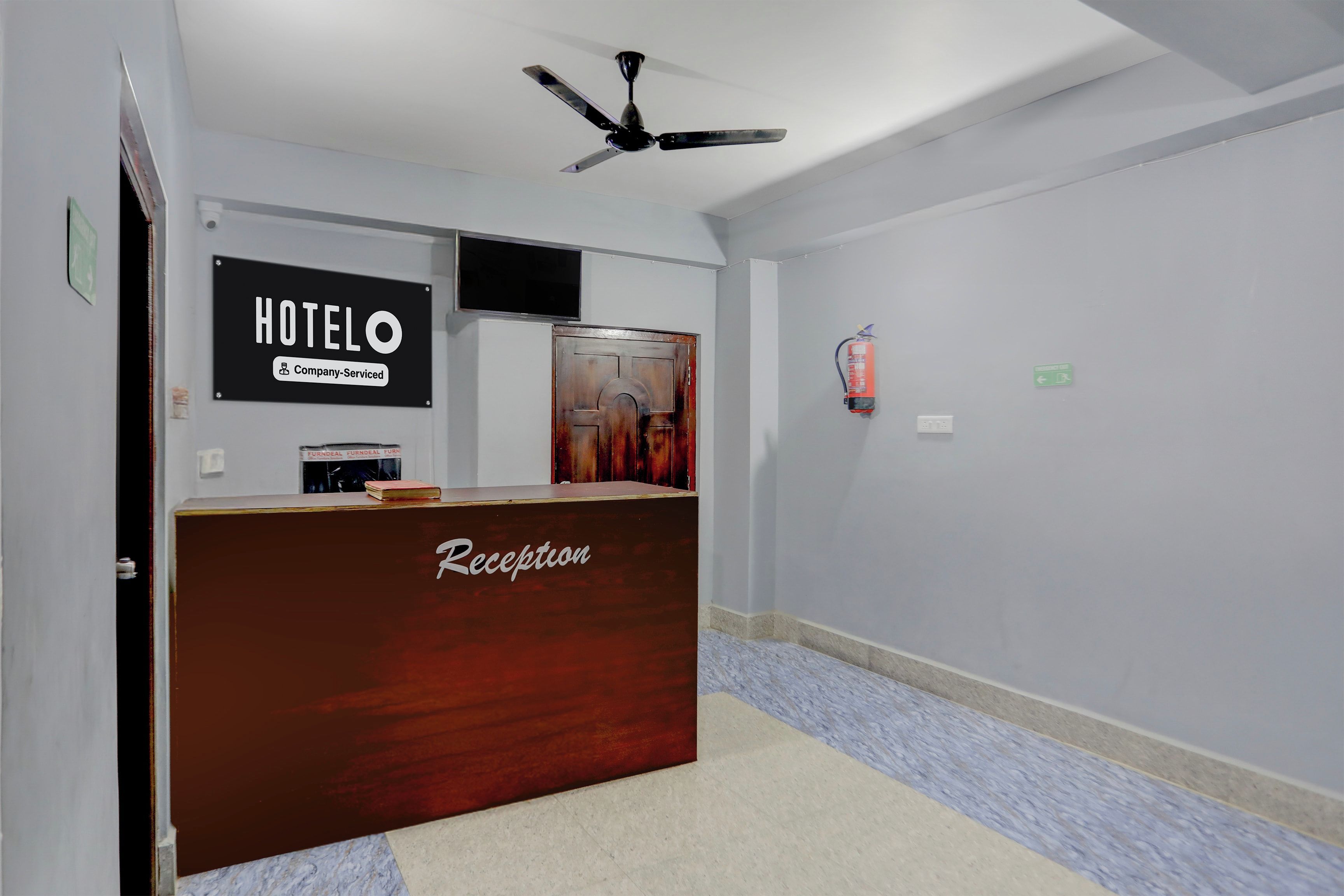 Una recepción de hotel con un cartel que indica que es un hotel de servicio empresarial. Hay un ventilador de techo arriba y un extintor en la pared.