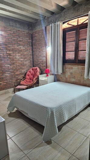 Una habitación con cama, una silla, una mesita de noche y un ventanal con cortinas.