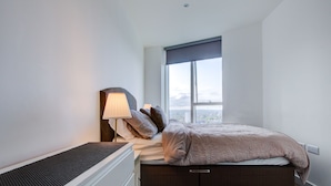 Een slaapkamer met een bed, nachtkastje, lamp en een raam met uitzicht op de stad.