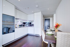 A modern kitchen with white cabinetry, a dining area with a table set for two, and a view into the living area. - Luxury City Apartment in Central London (London)