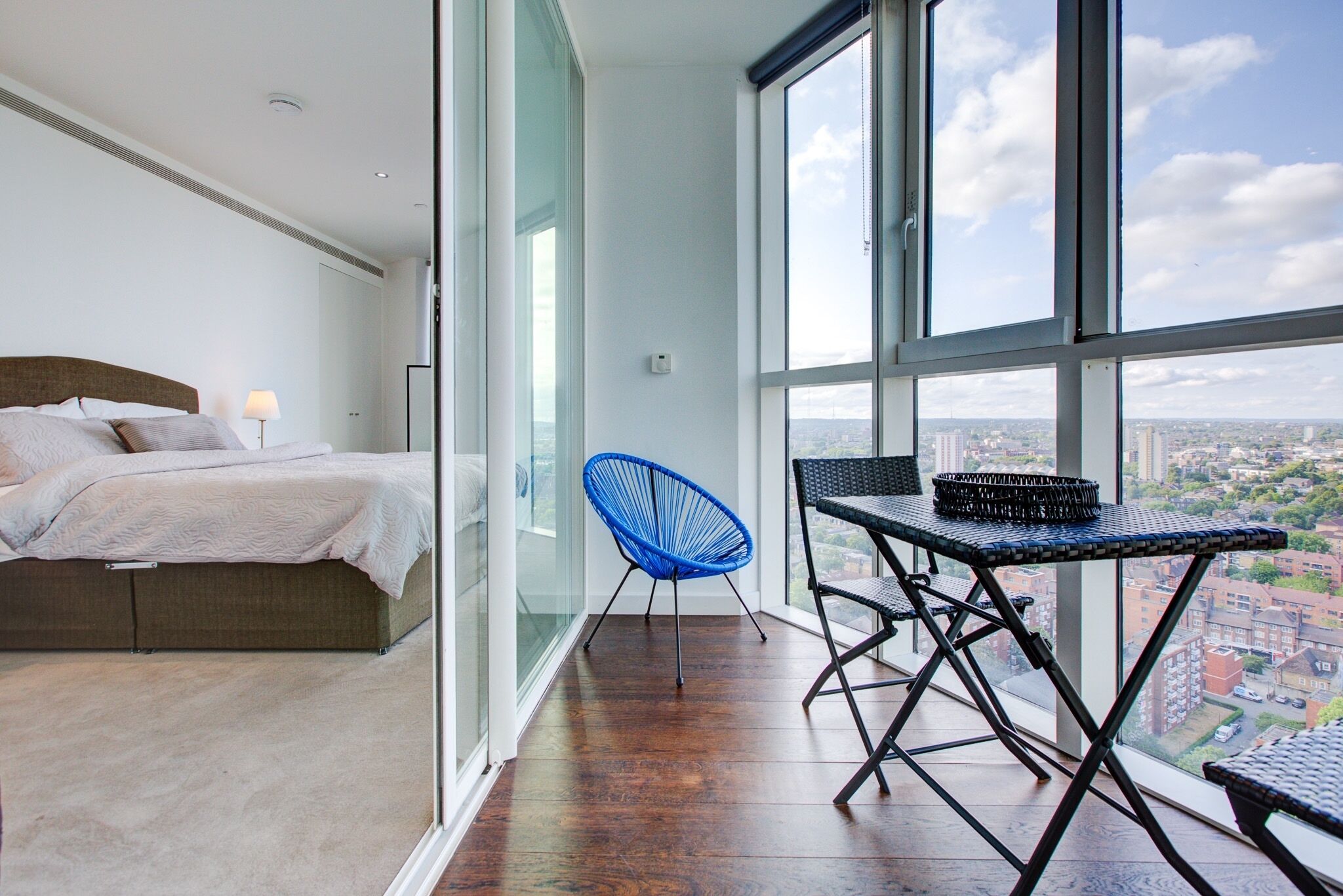 A bedroom with a bed and a nightstand, a balcony with a chair and a table, and a city view through large windows.