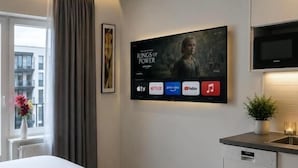 Ein Hotelzimmer mit Bett, Flachbildfernseher an der Wand, einer kleinen Küchenzeile inklusive Geschirrspüler und einem Fenster mit Vorhängen.