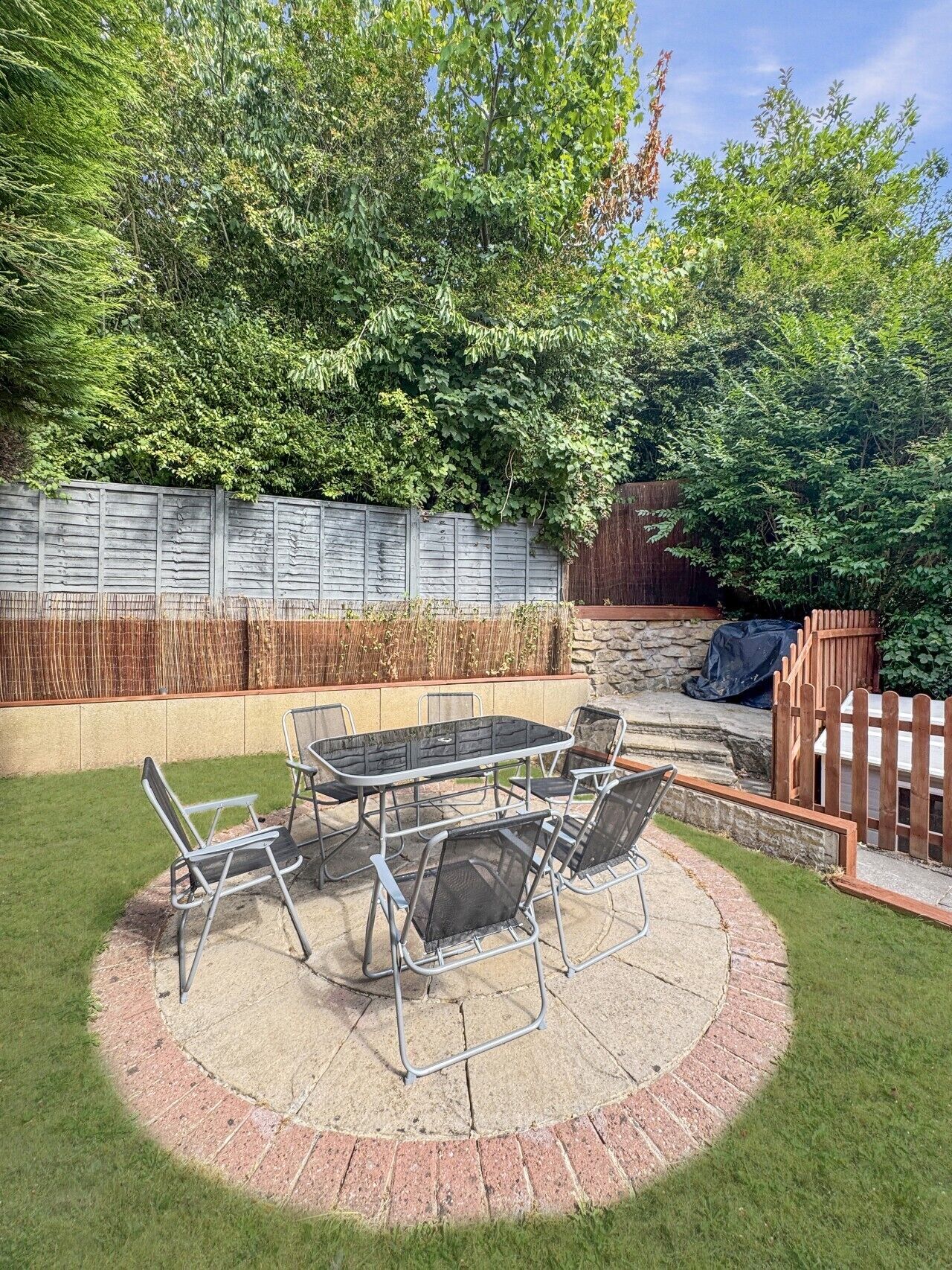 Une terrasse circulaire avec une table et des chaises, entourée d’une bordure en briques, située dans un jardin avec une clôture en bois et de la verdure.