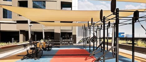 Une salle de sport en plein air, avec des appareils d’exercice, un tapis rouge et un bâtiment en arrière-plan.