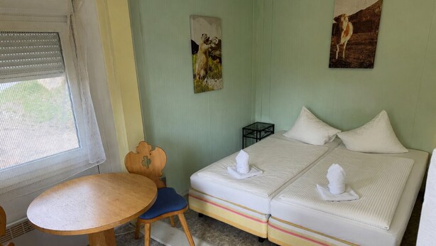 Une pièce avec un lit, une petite table et une chaise. Il y a une fenêtre avec des stores et deux tableaux accrochés au mur.