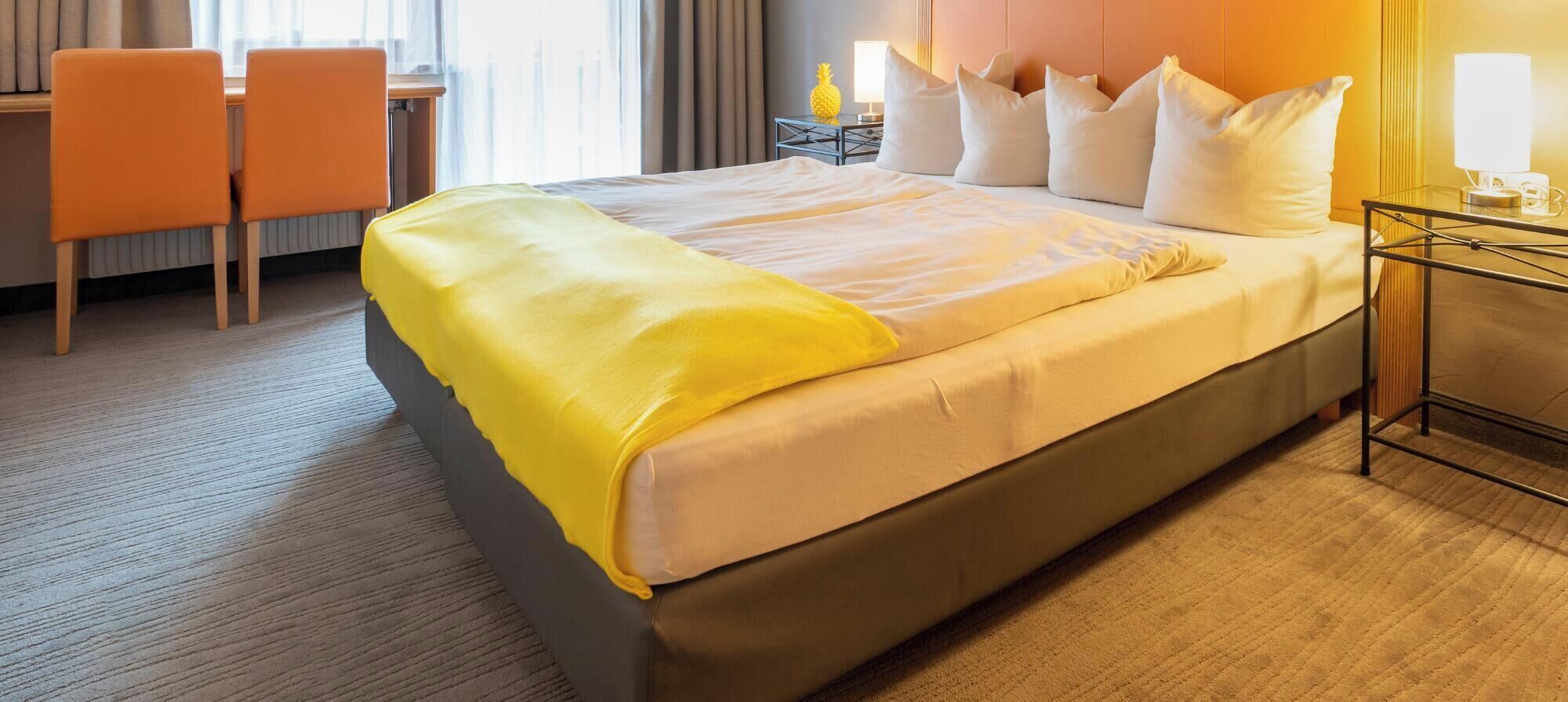 A hotel room with a bed, a desk, two chairs, and a nightstand with a lamp.