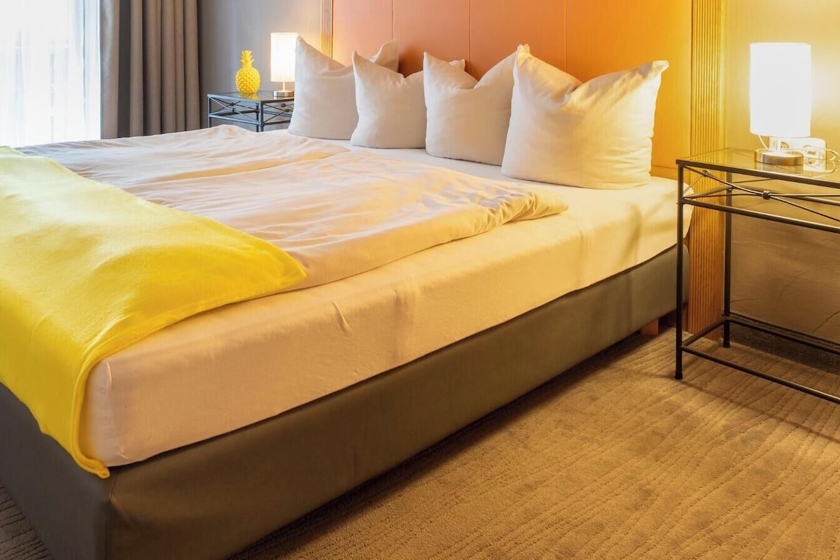 A hotel room with a bed, a desk, two chairs, and a nightstand with a lamp.
