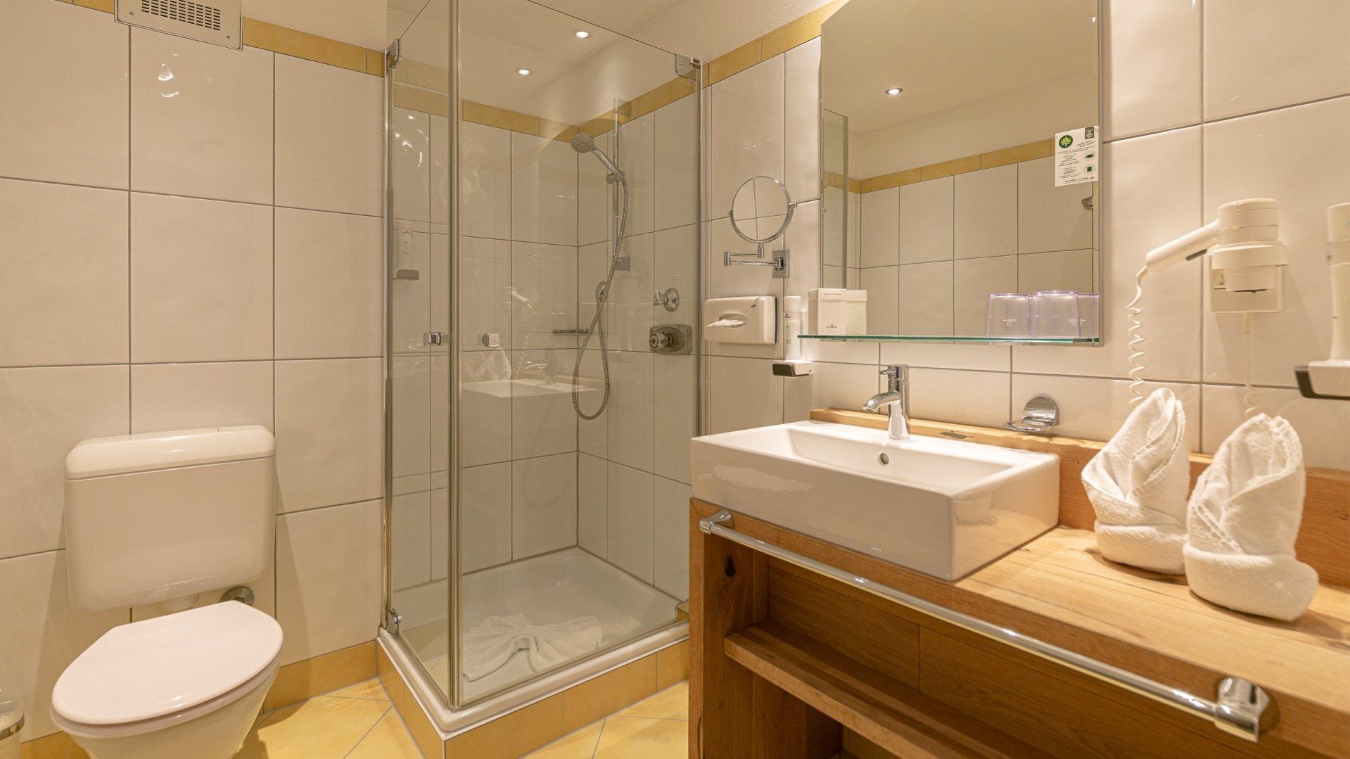 Un baño moderno con ducha envuelta en vidrio, inodoro blanco, lavabo con espejo y tocador de madera.