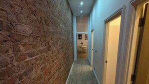 Ein Korridor mit einer Backsteinwand und Holzdielen. Rechts befinden sich Türen, und die Decke ist mit Einbaustrahlern versehen.