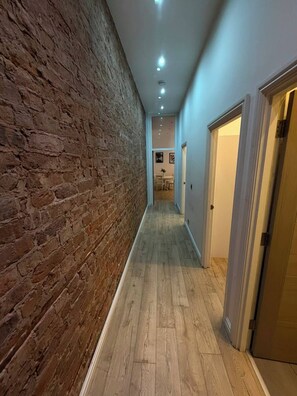Ein Korridor mit einer Backsteinwand und Holzdielen. Rechts befinden sich Türen, und die Decke ist mit Einbaustrahlern versehen.