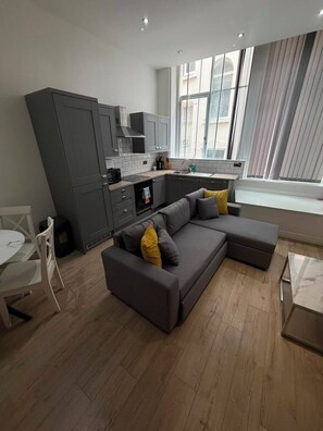 A modern living room with a grey sectional sofa, a dining table with chairs, and a kitchen area with grey cabinets and a window. - Sweeting Street Apartments (Liverpool)