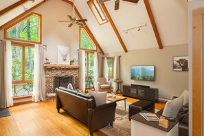 A spacious living room with a stone fireplace, leather sofas, a television, and large windows offering a view of the outdoors. - FirePit, Hot Tub, Dock + Lake "Get Your Creek On" (Swanton)