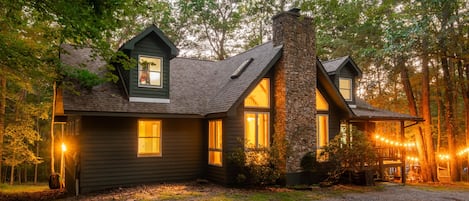 A cabin with a stone chimney, a porch with string lights, and a view of the surrounding forest.