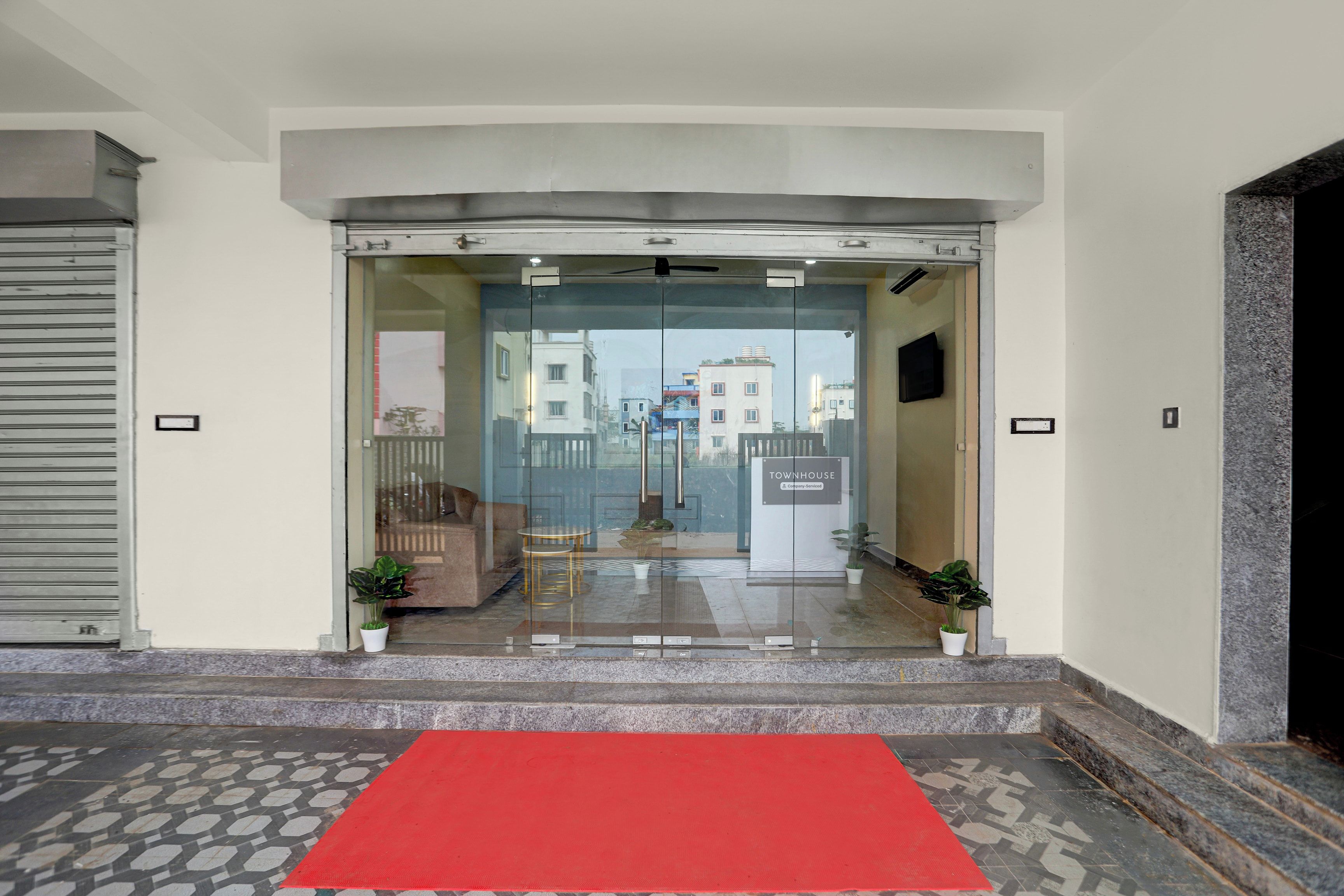 Entrada de hotel con alfombra roja, puertas de cristal y vista de la calle y edificios al fondo.