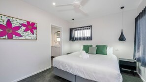 A bedroom with a bed, a ceiling fan, a window with curtains, a bedside table, and a wall-mounted lamp. - Shoreline Apartments - unit 13 (Bargara)