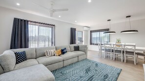 A modern living room with a grey sofa, patterned rug, and a dining area with a white table and chairs. - Shoreline Apartments - unit 13 (Bargara)