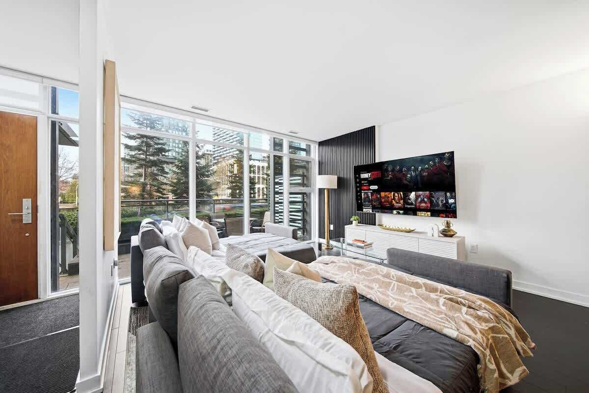 A modern living room with a sofa set, a flat-screen TV, and a large window offering a view of the outdoors.