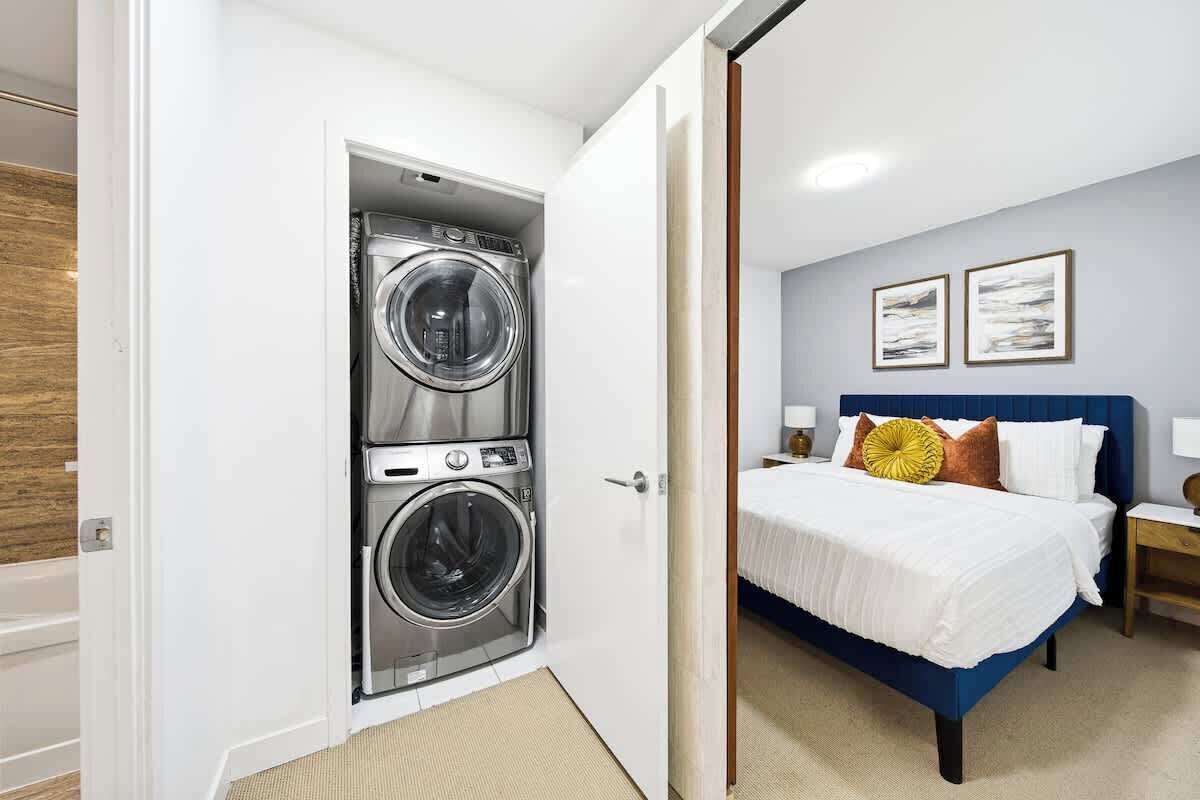 Hotellihuoneessa on sänky, yöpöytä, lamppu, seinälle ripustettu taideteos sekä ovi, joka johtaa pyykinpesutilaan, jossa on kaksi pesukonetta.