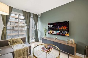 Un salon moderne doté d’un grand téléviseur à écran plat diffusant un film, d’un canapé, d’une table basse et offrant une vue sur un bâtiment à l’extérieur.