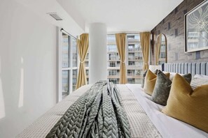 A hotel room with a large bed, multiple pillows, a view of a building through the window, and a modern design. - Spacious King Suite |CN Tower |Sleeps 5 w/Parking! (Toronto)