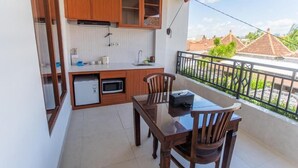 A compact kitchen with wooden cabinets, a refrigerator, and a microwave. There is a dining table with chairs and a small window overlooking a balcony with a view of buildings and greenery. - Pondok Bali 2 Homestay (Canggu)