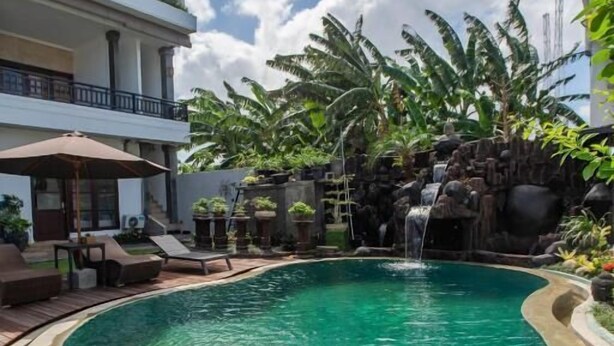 A swimming pool with a waterfall feature, surrounded by lounge chairs and tropical plants. - Pondok Bali 2 Homestay (Canggu)