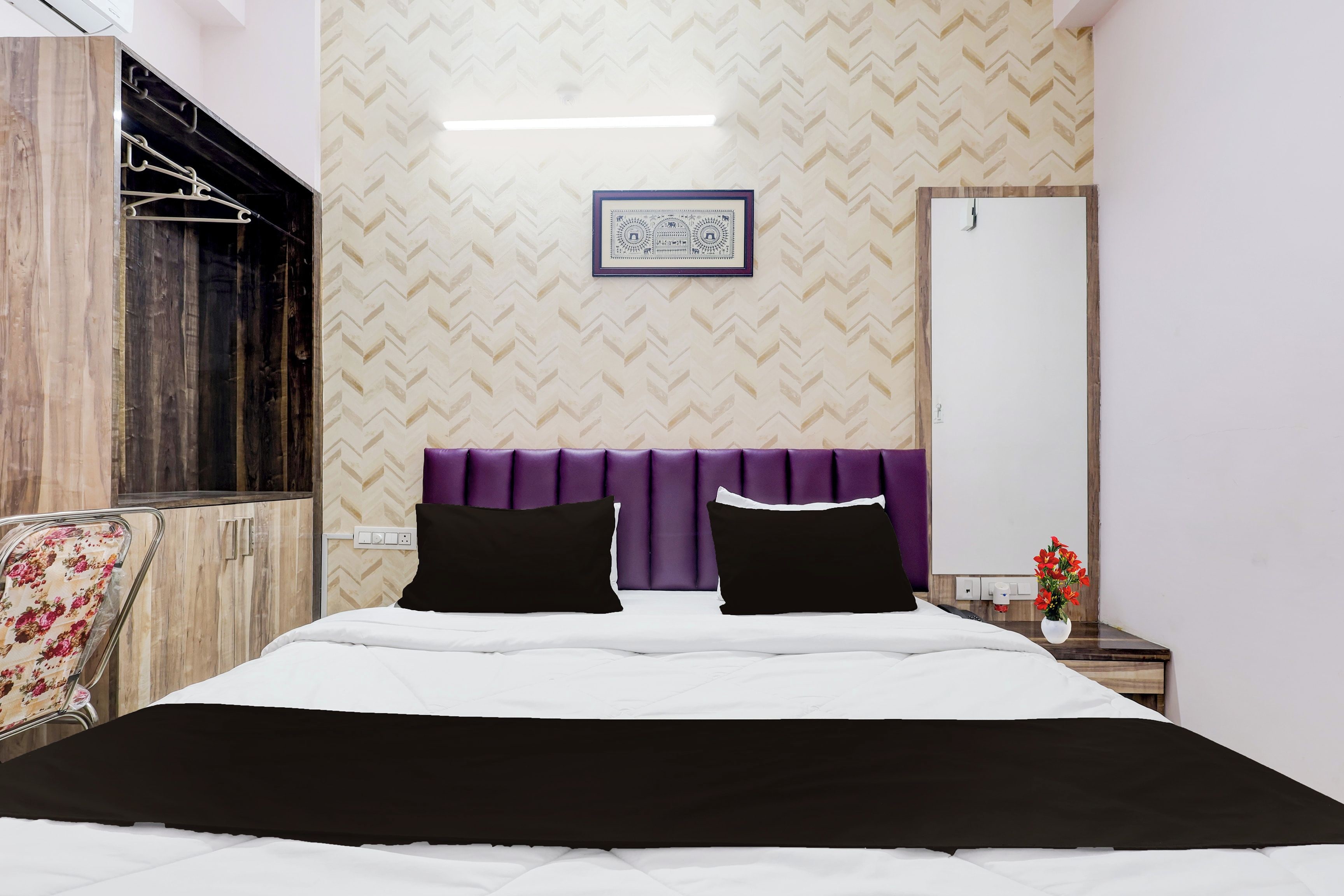 A hotel room with a bed, a mirror, a wall-mounted picture, and a small vase with flowers.