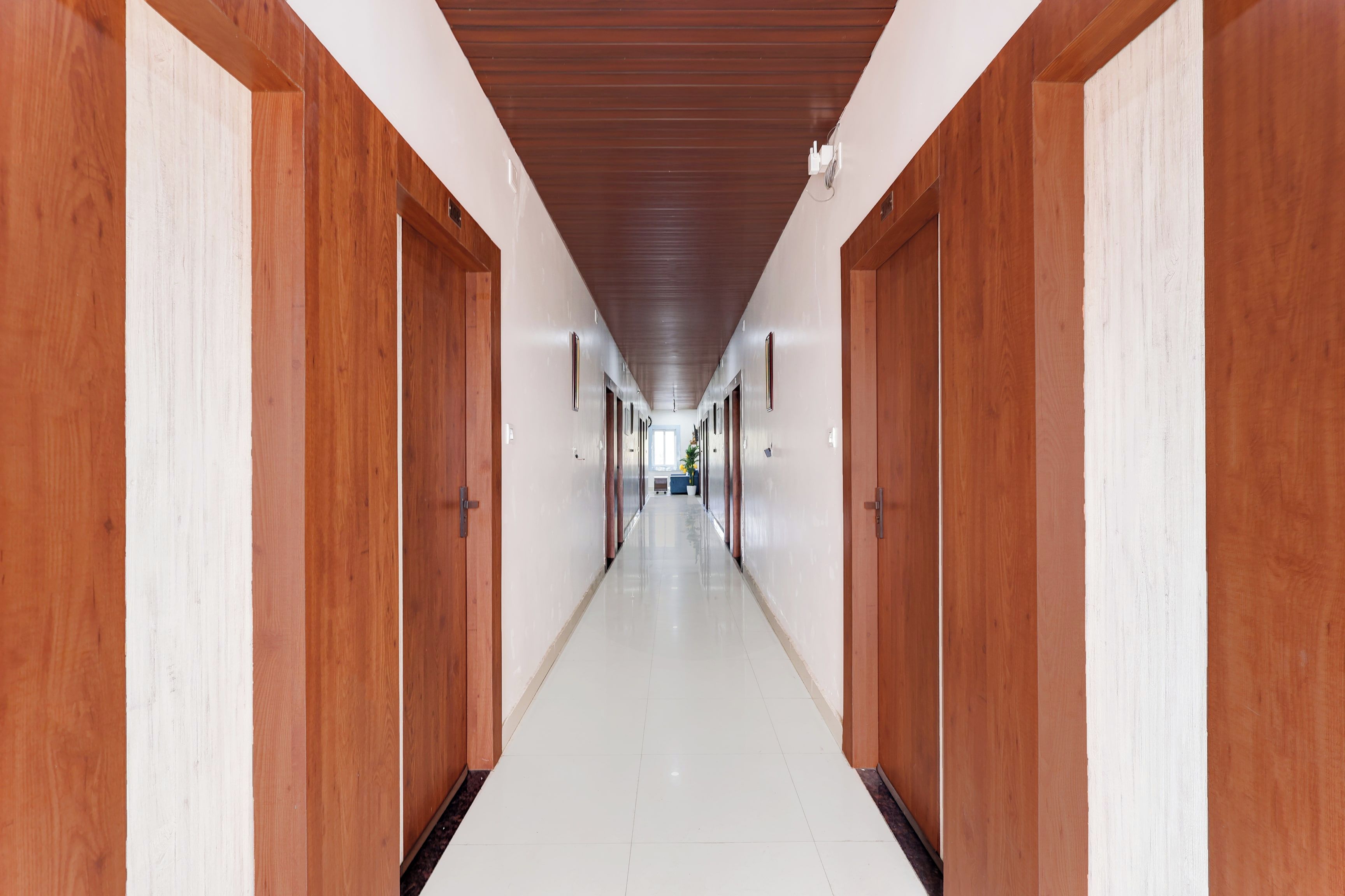 Un largo y estrecho pasillo con puertas de madera a ambos lados y piso de baldosas blancas.