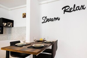 Ein Essbereich mit einem Tisch für zwei Personen, einer Wand mit der Aufschrift 'Relax Dream' in schwarzer Schrift und einem kleinen Regal mit dekorativen Gegenständen.