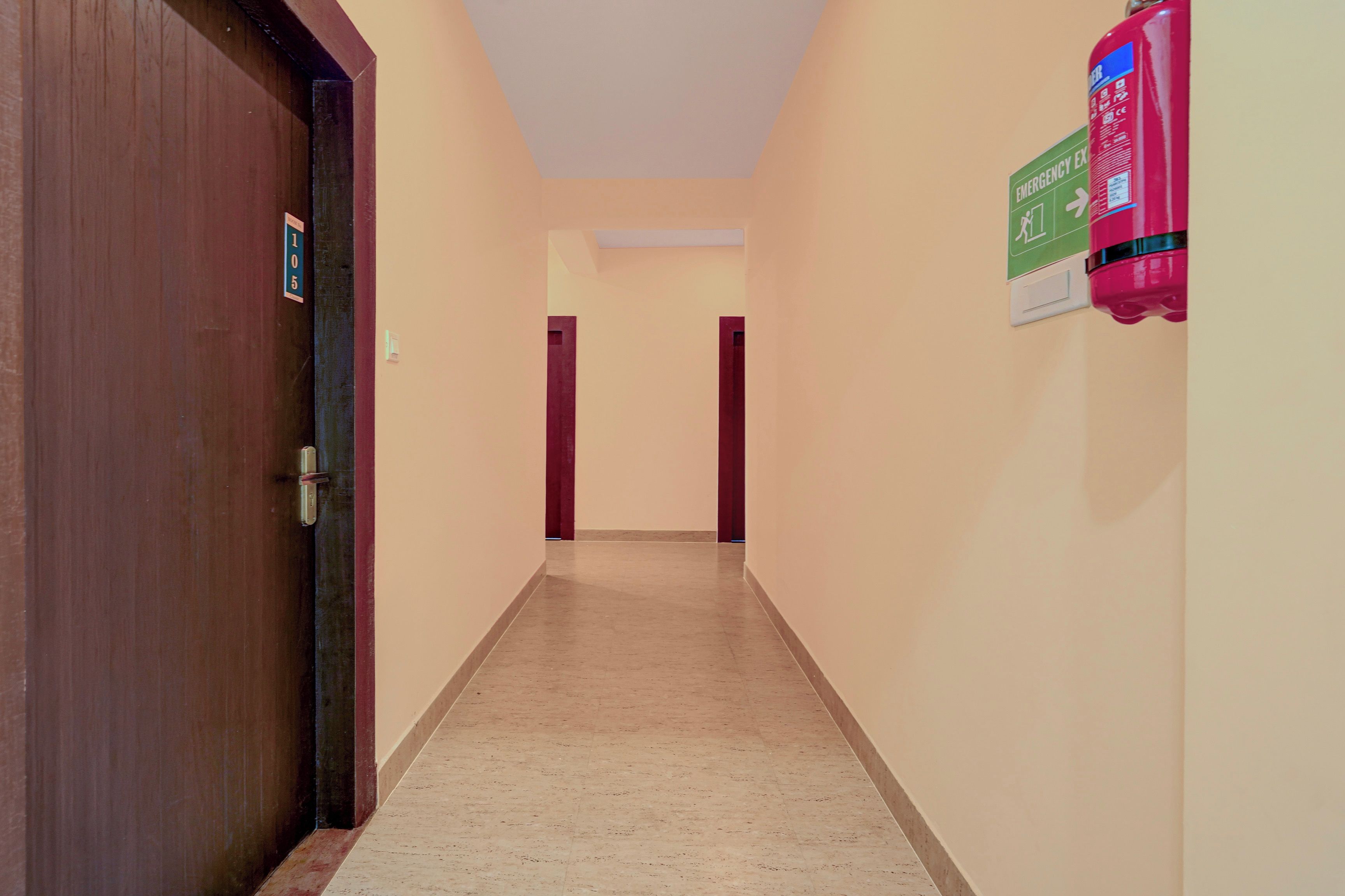 Un pasillo con una puerta de madera a la izquierda, un extintor a la derecha y un letrero de salida de emergencia verde en la pared.