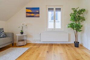 Ein Wohnzimmer mit grauem Sofa, einem kleinen runden Tisch mit orchideenbewzter Pflanze, einer Heizung, einem Fenster mit Jalousien und einem großen Topfbaum.