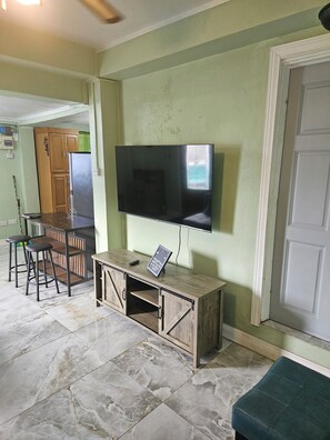 Ein Wohnzimmer mit Flachbildfernseher an einem Holzkasten, eine Essecke mit Tisch und Stühlen sowie eine Küchenzeile mit Kühlschrank.