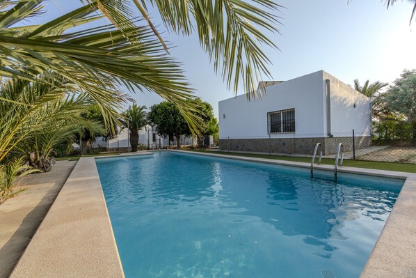 Una piscina con agua azul cristalina, rodeada de palmeras y un edificio blanco con una ventana.