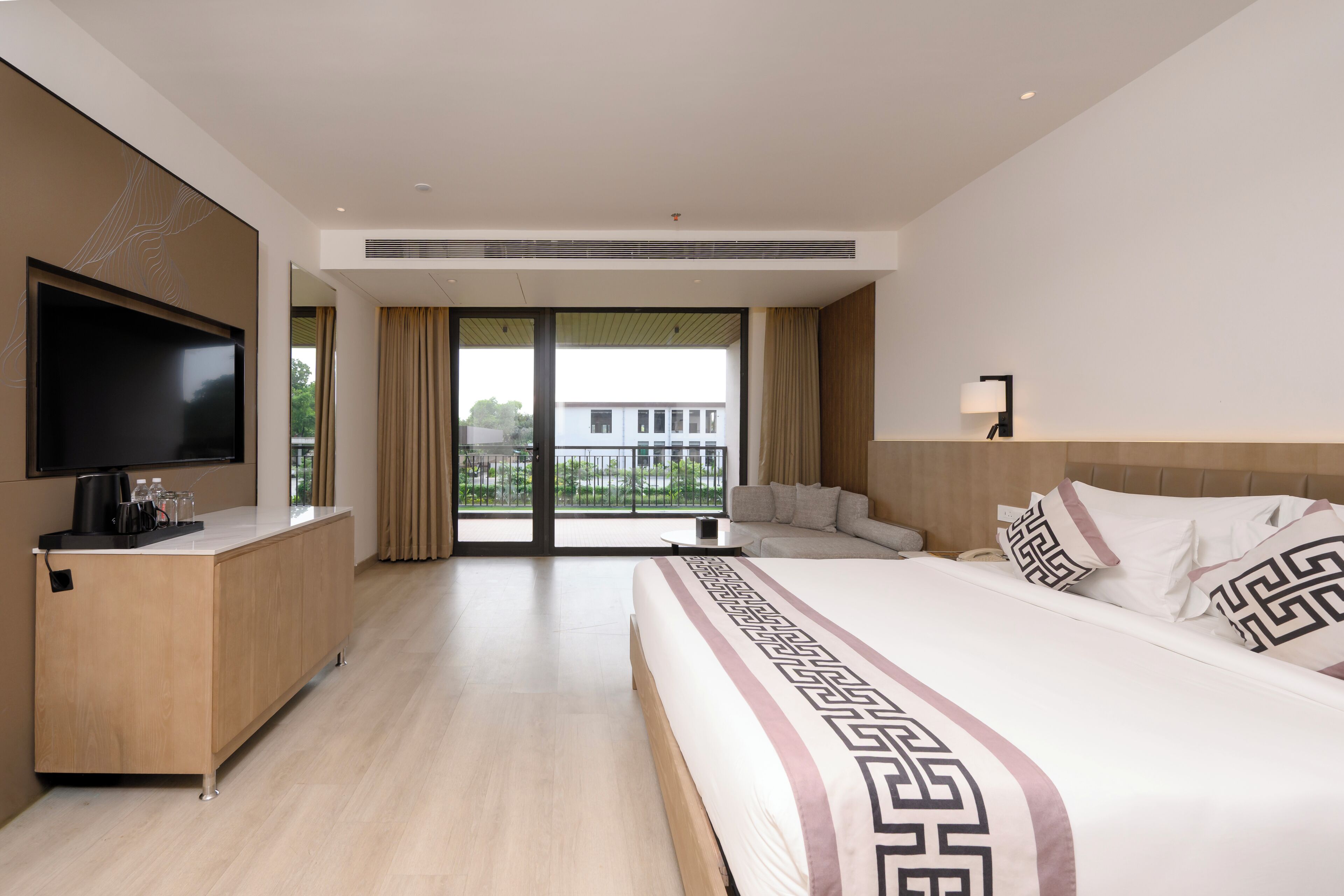 Ein modernes Hotelzimmer mit einem großen Bett, einem Flachbildfernseher, einem Schreibtisch mit Kaffeemaschine und einem Balkon mit Blick auf Gebäude.