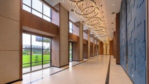 A spacious corridor with large windows, a modern chandelier, and a view of a building and greenery outside. - NANDGAON KASHI RESORT AND SPA (Varanasi)