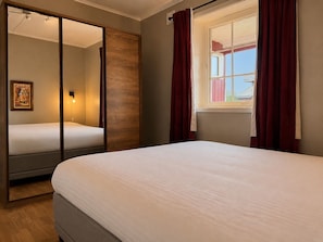 Un dormitorio con cama, espejo, ventana con cortinas y una lámpara fijada a la pared.