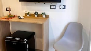 Una habitación con un televisor de pantalla plana montado en la pared, un escritorio de madera con un pequeño aparato negro, una silla gris y una planta en maceta.