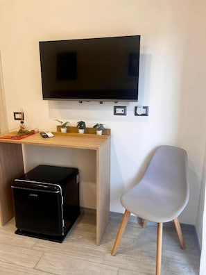 A room with a flat-screen TV mounted on the wall, a wooden desk with a small black appliance, a grey chair, and a potted plant.
