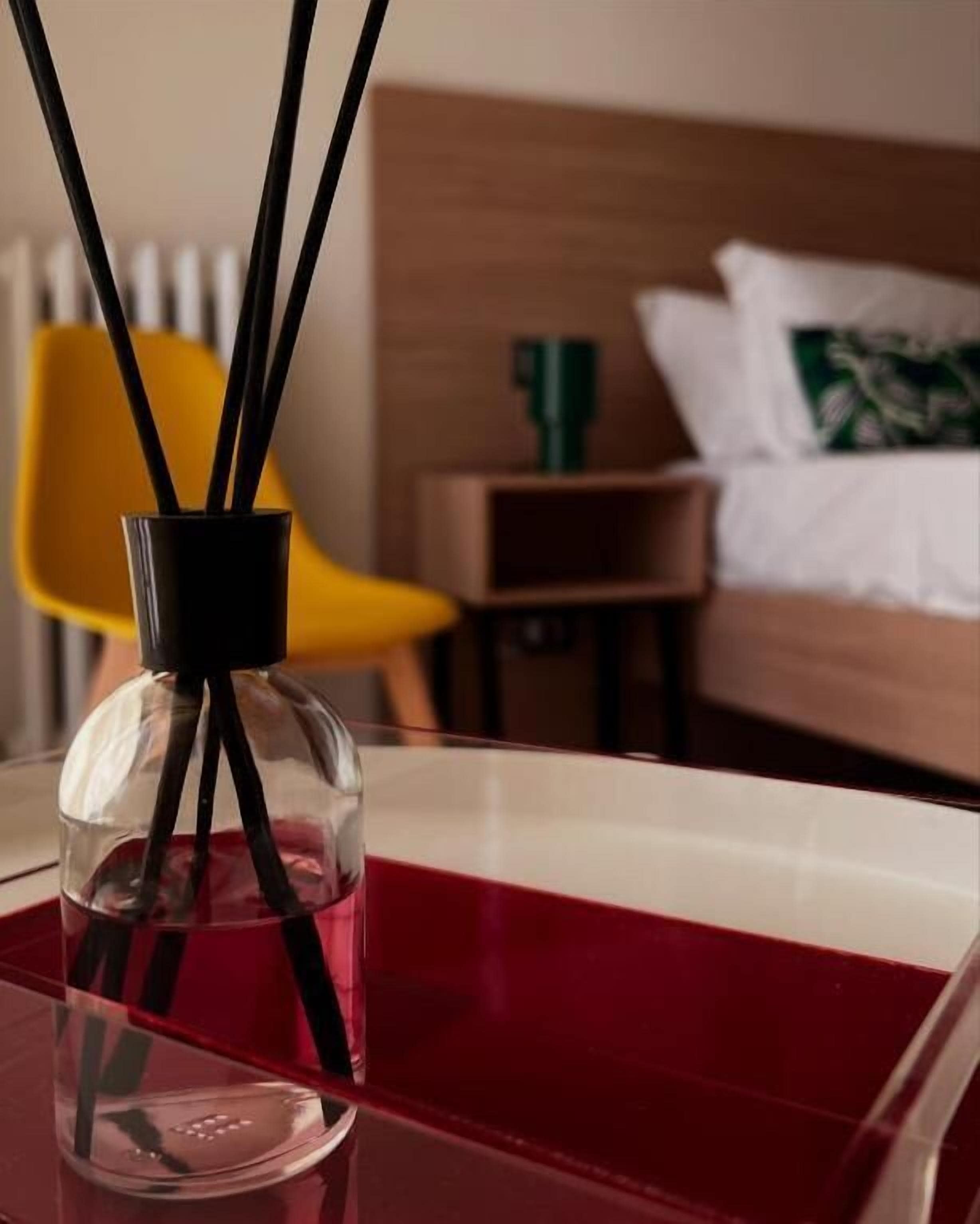 Ein Hotelzimmer mit einem Bett, einem Nachttisch, einer roten Ablage und einer durchsichtigen Flasche mit schwarzen Stäbchen.