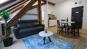 Ett modernt vardagsrum med en svart lädersoffa, ett runt, vitt soffbord och en blå, mönstrad matta. Det finns en matplats med ett vitt bord och svarta stolar, och ett kök syns i bakgrunden.