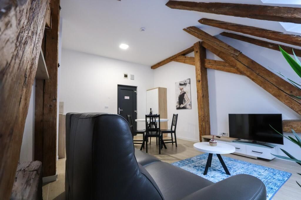Un salon moderne avec un canapé en cuir noir, une table à manger avec des chaises, une petite table blanche sur laquelle se trouve un téléviseur, et une poutre en bois au plafond.