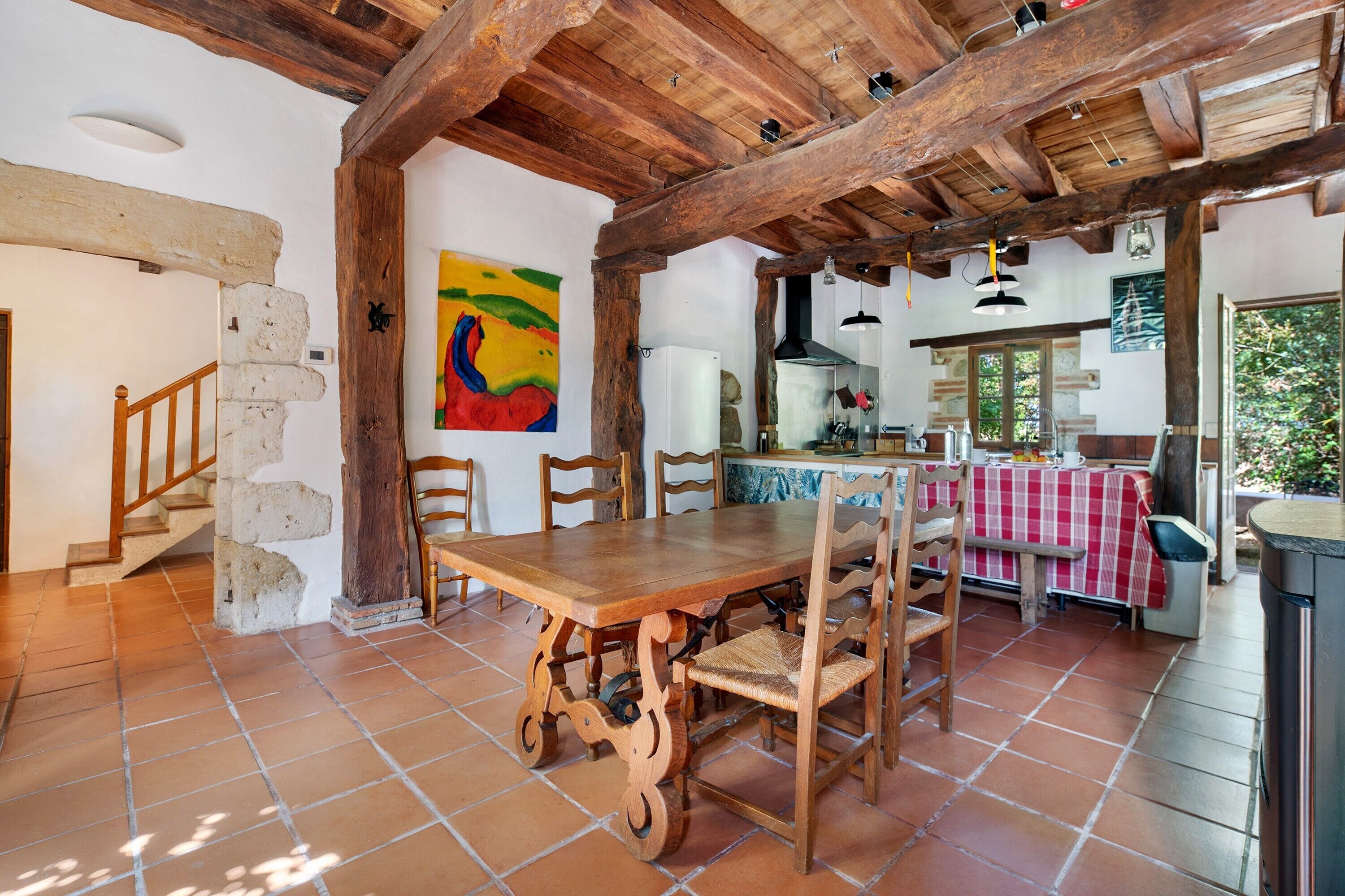 Un interior rústico con una mesa de comedor de madera, sillas, una zona de cocina y una escalera.