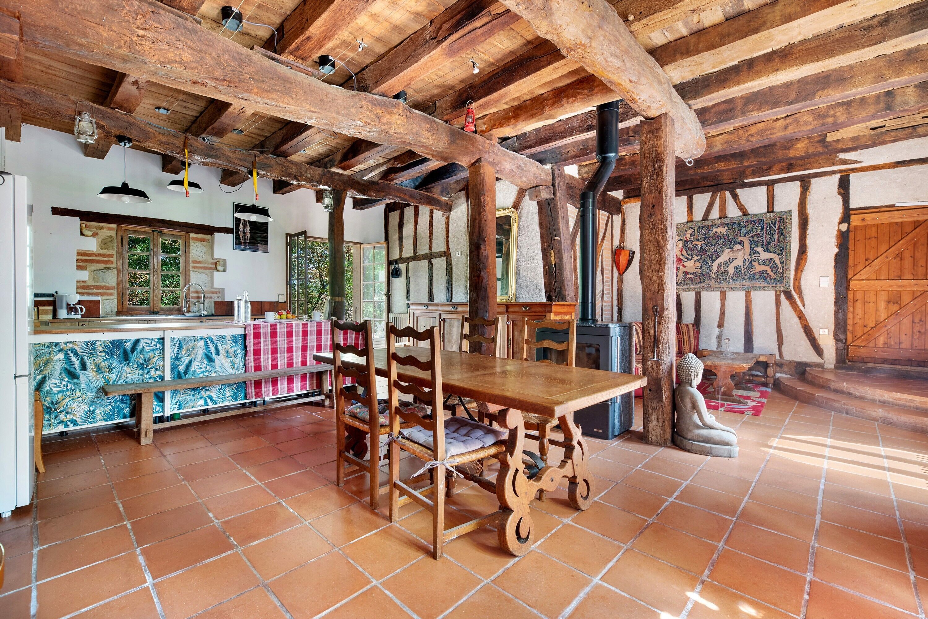 A rustic interior with exposed wooden beams, a tiled floor, a dining area with wooden chairs, a kitchen with a tiled backsplash, and a large wooden door.