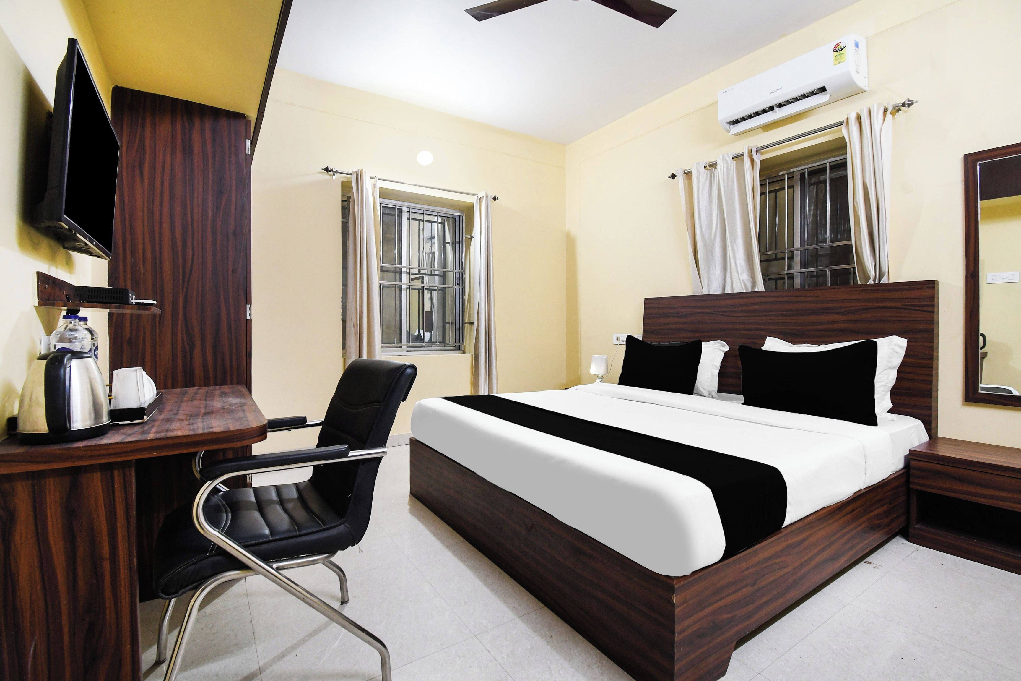 A hotel room with a bed, a desk with a chair, a television, an air conditioning unit, and a window with curtains.